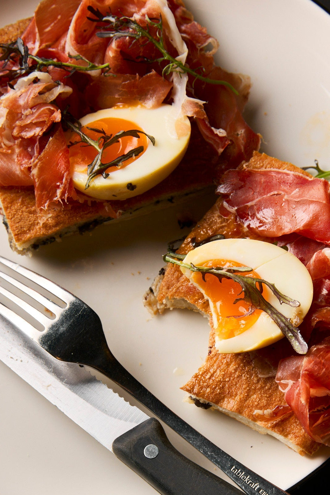 Plated dish with bread topped with prosciutto, egg, and herbs, accompanied by a knife and glass of iced coffee.