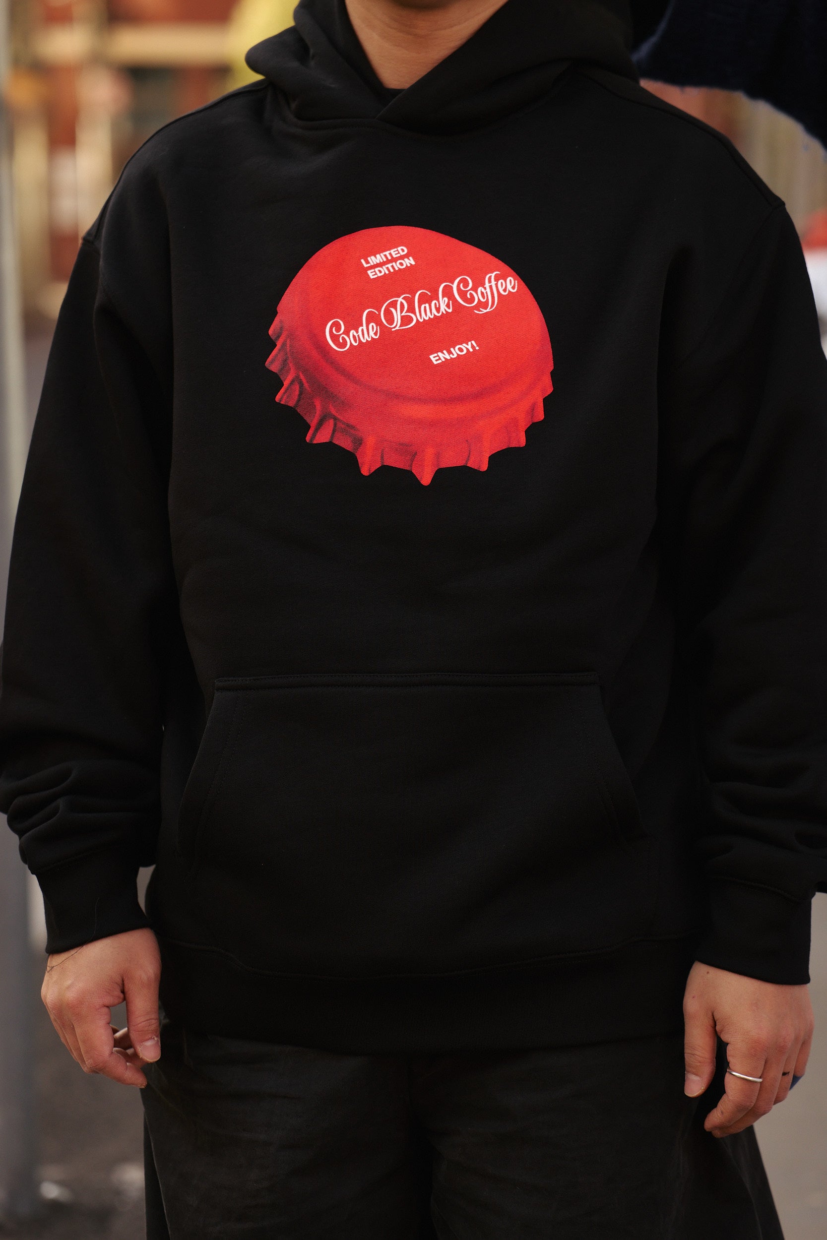 A person wearing a black pullover hoodie with a large red bottle-cap graphic on the chest featuring the text 'Code Black Coffee' and 'LIMITED EDITION'.
