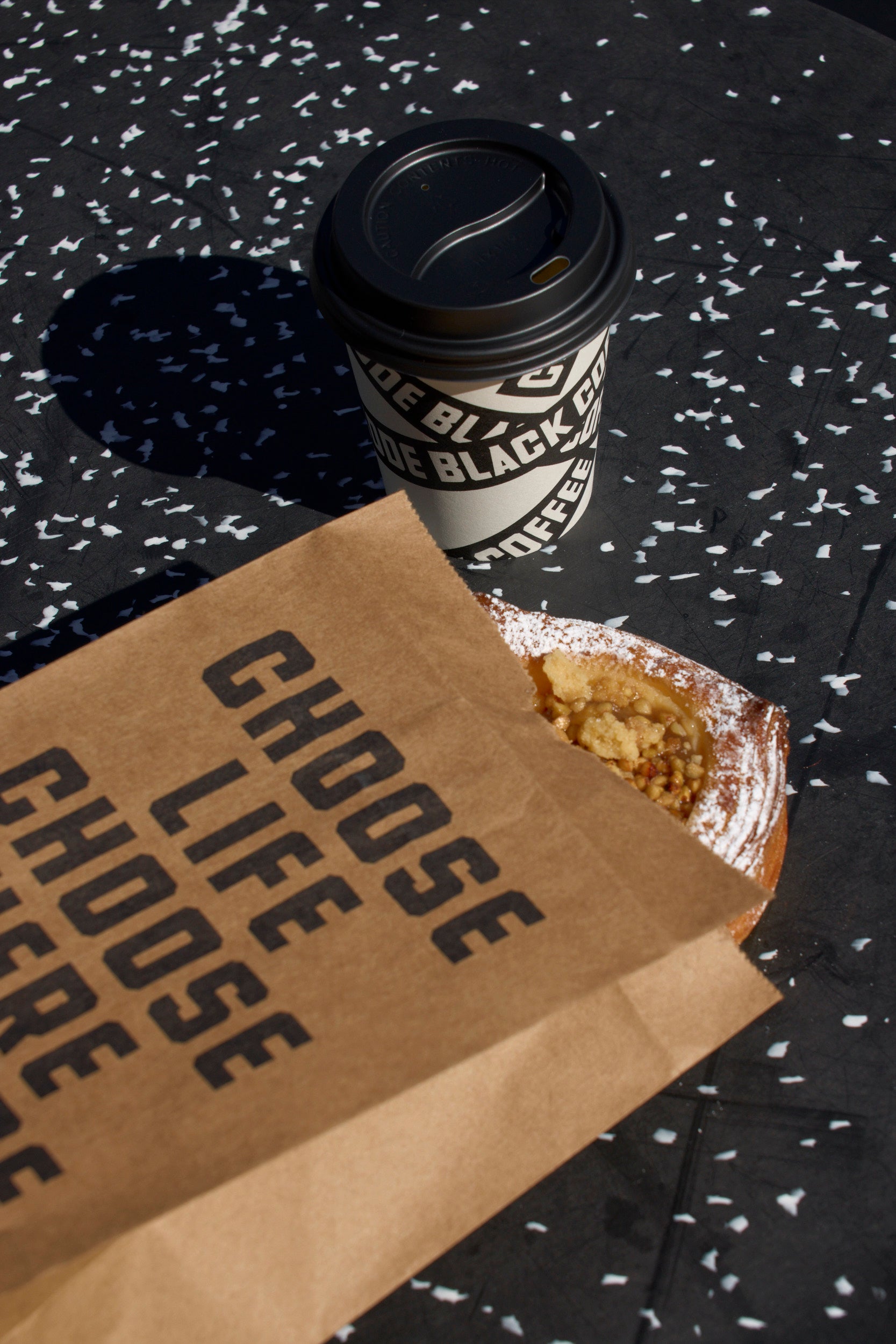 Code Black Coffee cup, brown paper bag with 'CHOOSE LIFE' text, and snack on a black speckled surface